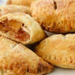 A plate of golden-brown hand pies, some with sugar sprinkled on top. One hand pie is cut in half, revealing a filling of diced apples and caramelized sugar. The edges of the pastries are crimped, giving them a rustic look.