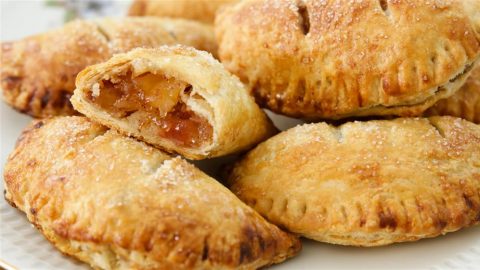 A plate of golden-brown hand pies, some with sugar sprinkled on top. One hand pie is cut in half, revealing a filling of diced apples and caramelized sugar. The edges of the pastries are crimped, giving them a rustic look.