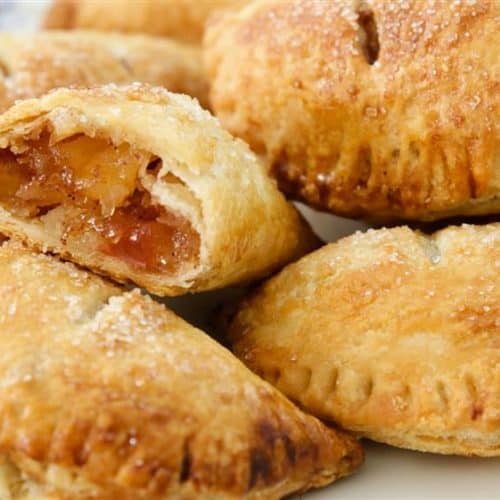 A plate of golden-brown hand pies, some with sugar sprinkled on top. One hand pie is cut in half, revealing a filling of diced apples and caramelized sugar. The edges of the pastries are crimped, giving them a rustic look.