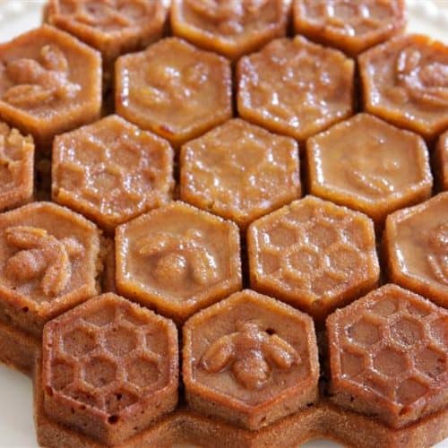 A cake with a honeycomb design, composed of hexagonal pieces. Each hexagon is decorated with detailed bee and honeycomb patterns, giving the cake an intricate, beehive-like appearance. The cake sits on a white plate.