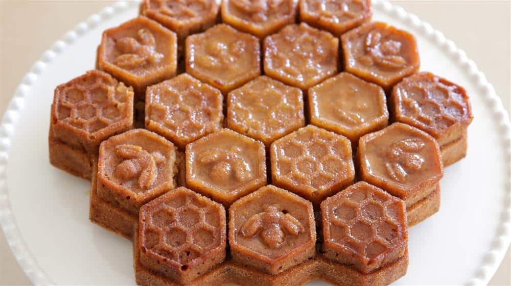 A cake with a honeycomb design, composed of hexagonal pieces. Each hexagon is decorated with detailed bee and honeycomb patterns, giving the cake an intricate, beehive-like appearance. The cake sits on a white plate.