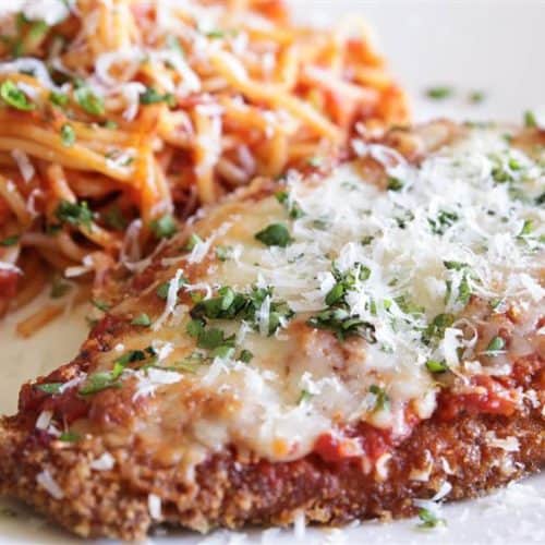 A plate with breaded chicken parmesan topped with melted cheese and marinara sauce, garnished with chopped parsley, next to a portion of spaghetti mixed with tomato sauce, sprinkled with grated cheese.