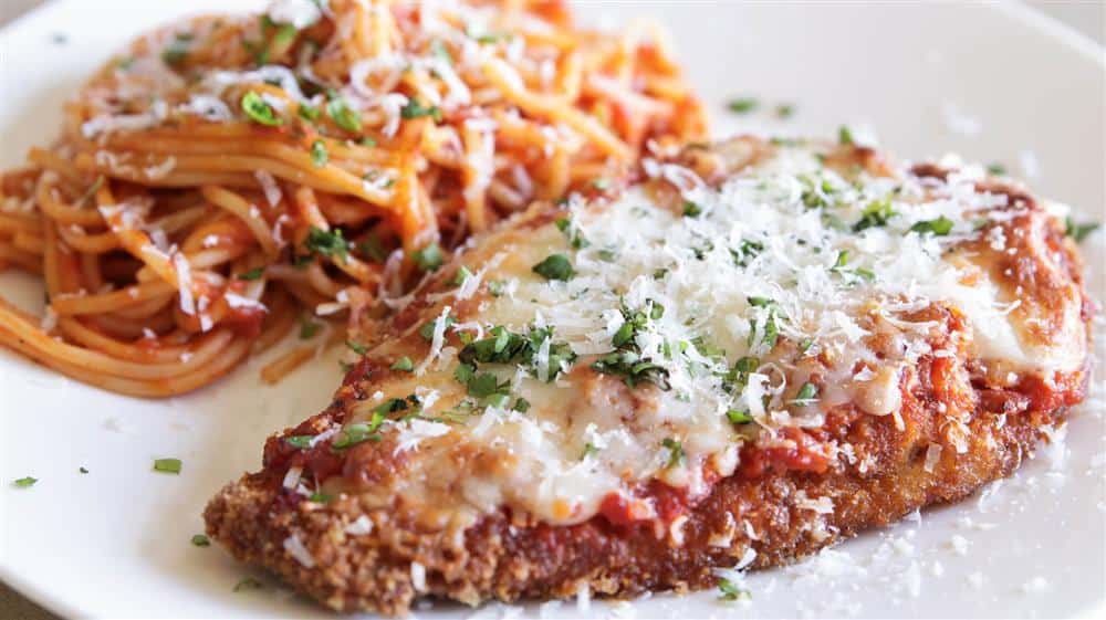 A plate with breaded chicken parmesan topped with melted cheese and marinara sauce, garnished with chopped parsley, next to a portion of spaghetti mixed with tomato sauce, sprinkled with grated cheese.