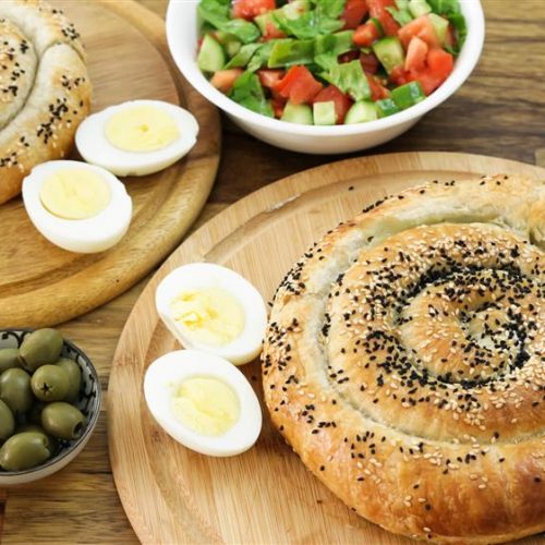 A wooden table displays two spiral-shaped pastries sprinkled with sesame and nigella seeds, one on each of two separate wooden boards. The boards also hold halved boiled eggs. A bowl with a chopped salad and another bowl with green olives complete the setting.