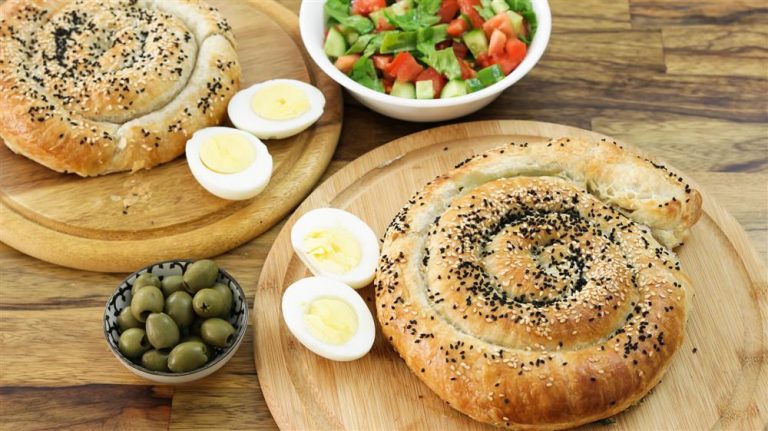 A wooden table displays two spiral-shaped pastries sprinkled with sesame and nigella seeds, one on each of two separate wooden boards. The boards also hold halved boiled eggs. A bowl with a chopped salad and another bowl with green olives complete the setting.