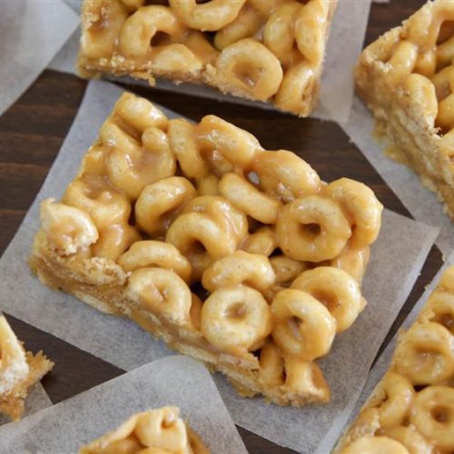 Rectangular cereal treats made with O-shaped cereal and a sticky, caramel-colored coating are arranged on parchment paper. The treats have a shiny appearance, indicating they may contain a sugary or honey-based binder.