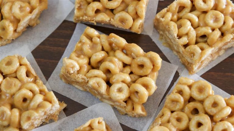 Rectangular cereal treats made with O-shaped cereal and a sticky, caramel-colored coating are arranged on parchment paper. The treats have a shiny appearance, indicating they may contain a sugary or honey-based binder.