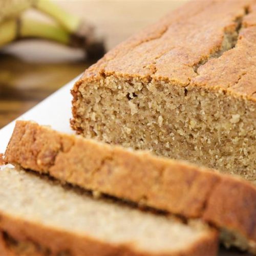 A close-up image of freshly baked banana bread on a white plate, with two slices cut from the loaf. The bread has a golden-brown crust and a moist interior. Ripe bananas are visible in the background.