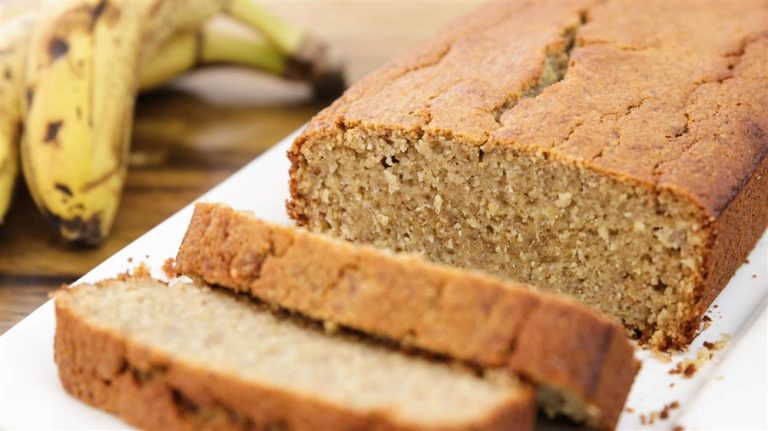 A close-up image of freshly baked banana bread on a white plate, with two slices cut from the loaf. The bread has a golden-brown crust and a moist interior. Ripe bananas are visible in the background.