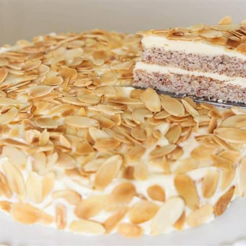 A round almond cake garnished with almond slices sits on a white cake stand. A slice is lifted from the cake, revealing layers of almond sponge cake and creamy filling. The cake is topped and adorned on the sides with toasted almond slices.