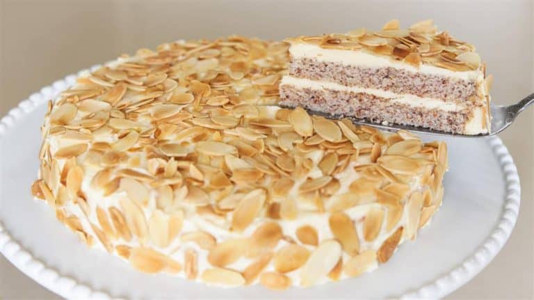 A round almond cake garnished with almond slices sits on a white cake stand. A slice is lifted from the cake, revealing layers of almond sponge cake and creamy filling. The cake is topped and adorned on the sides with toasted almond slices.