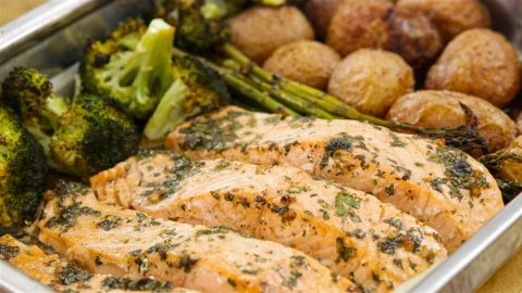 A close-up photo of a baked salmon dish garnished with herbs. The salmon fillets are accompanied by roasted broccoli, asparagus, and baby potatoes, all served in a metal tray. The food appears freshly cooked and ready to serve.