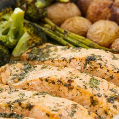 A close-up photo of a baked salmon dish garnished with herbs. The salmon fillets are accompanied by roasted broccoli, asparagus, and baby potatoes, all served in a metal tray. The food appears freshly cooked and ready to serve.