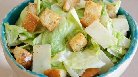 A close-up of a Caesar salad in a blue bowl, featuring crisp romaine lettuce, crunchy croutons, and shavings of parmesan cheese. The salad is lightly dressed, giving it a fresh and appetizing appearance.