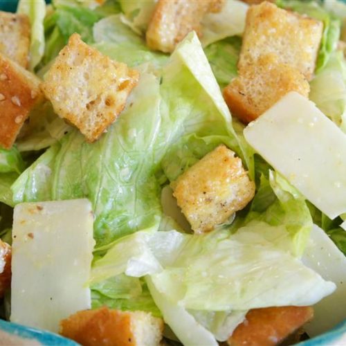 A close-up of a Caesar salad in a blue bowl, featuring crisp romaine lettuce, crunchy croutons, and shavings of parmesan cheese. The salad is lightly dressed, giving it a fresh and appetizing appearance.