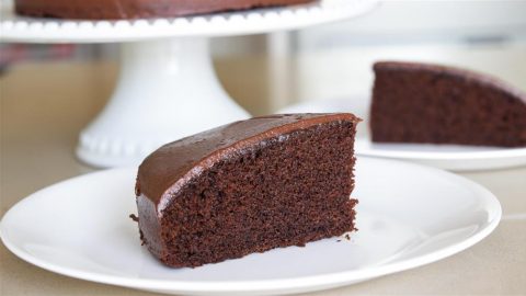 A slice of chocolate cake with smooth chocolate frosting is placed on a white plate. In the background, the remaining cake on a cake stand and another slice on a separate plate are visible. The cake appears moist and rich in texture.