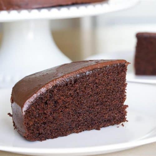 A slice of chocolate cake with smooth chocolate frosting is placed on a white plate. In the background, the remaining cake on a cake stand and another slice on a separate plate are visible. The cake appears moist and rich in texture.