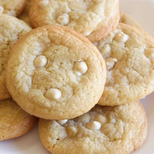 A close-up of a plate with several white chocolate chip cookies. The cookies are golden brown with visible white chocolate chips, and they are arranged in a slightly overlapping pattern on a plain white plate.