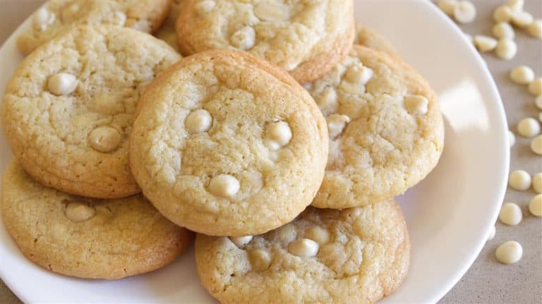 A close-up of a plate with several white chocolate chip cookies. The cookies are golden brown with visible white chocolate chips, and they are arranged in a slightly overlapping pattern on a plain white plate.