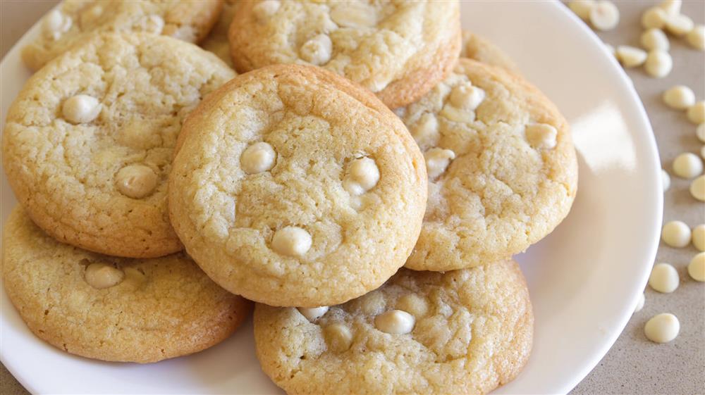 A close-up of a plate with several white chocolate chip cookies. The cookies are golden brown with visible white chocolate chips, and they are arranged in a slightly overlapping pattern on a plain white plate.