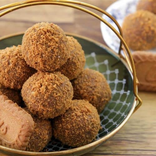 A basket filled with round Biscoff truffles, topped with a dusting of crumbs, placed on a green tray. Nearby, a plate holds a few more truffles and a rectangular Biscoff cookie, creating a warm, inviting dessert presentation.