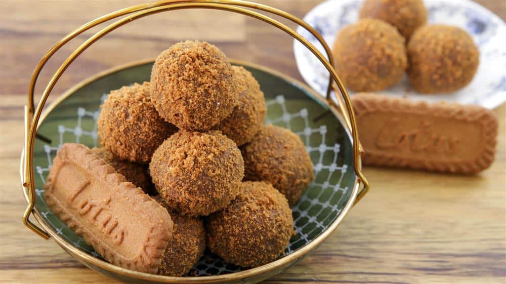 A basket filled with round Biscoff truffles, topped with a dusting of crumbs, placed on a green tray. Nearby, a plate holds a few more truffles and a rectangular Biscoff cookie, creating a warm, inviting dessert presentation.