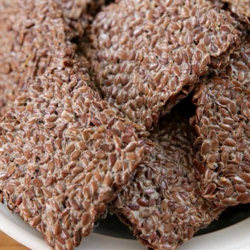 A close-up of a white bowl filled with flaxseed crackers. The crackers are rectangular and have a textured, grainy surface, showcasing the individual flaxseeds. The background is a wooden surface.