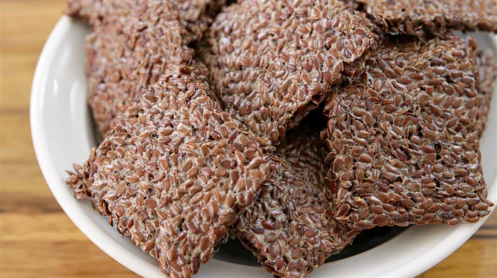 A close-up of a white bowl filled with flaxseed crackers. The crackers are rectangular and have a textured, grainy surface, showcasing the individual flaxseeds. The background is a wooden surface.