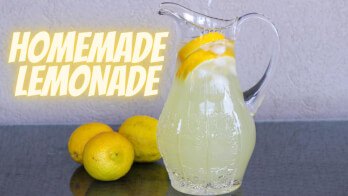 A clear pitcher filled with homemade lemonade, garnished with lemon slices and ice cubes, is placed on a countertop with a few whole lemons beside it. The text "HOMEMADE LEMONADE" is displayed in bold yellow letters to the left of the pitcher.