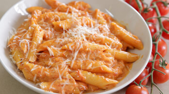 A white plate filled with penne pasta coated in a creamy tomato sauce and topped with grated cheese. Fresh cherry tomatoes on the vine are placed beside the plate.