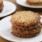 A close-up of a plate with a stack of oatmeal cookies on a wooden surface. Two additional plates with more cookies are blurred in the background. The cookies appear golden brown and crispy. The plates are white with a subtle floral design.