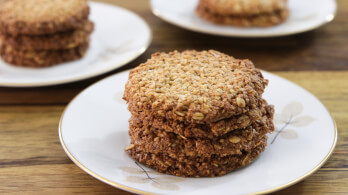 A close-up of a plate with a stack of oatmeal cookies on a wooden surface. Two additional plates with more cookies are blurred in the background. The cookies appear golden brown and crispy. The plates are white with a subtle floral design.