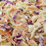 A close-up view of a bowl filled with coleslaw. The coleslaw is made up of shredded cabbage, both green and purple, as well as thin strips of carrots, all mixed together with a creamy dressing. The dish looks fresh and colorful.