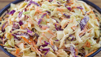 A close-up view of a bowl filled with coleslaw. The coleslaw is made up of shredded cabbage, both green and purple, as well as thin strips of carrots, all mixed together with a creamy dressing. The dish looks fresh and colorful.