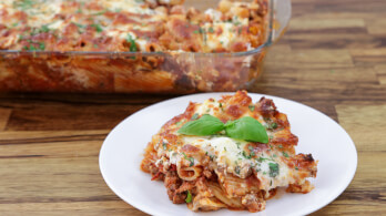 A serving of baked ziti with melted cheese and basil on top is placed on a white plate. In the background, a glass baking dish is filled with more baked ziti. The dish is set on a wooden table.