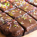 A close-up of sliced chocolate brownies topped with a glossy layer of frosting and colorful sprinkles. The brownies are neatly arranged on a wooden surface, with each piece evenly cut.