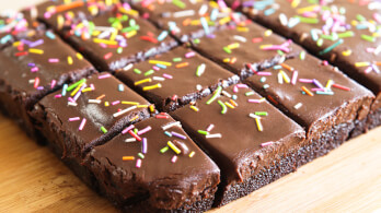 A close-up of sliced chocolate brownies topped with a glossy layer of frosting and colorful sprinkles. The brownies are neatly arranged on a wooden surface, with each piece evenly cut.