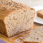 A freshly baked loaf of whole grain bread is sliced, with one slice placed on a wooden cutting board and another slice on a white plate, spread with a light layer of peanut butter. The bread is sprinkled with various seeds.