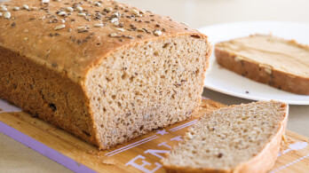 A freshly baked loaf of whole grain bread is sliced, with one slice placed on a wooden cutting board and another slice on a white plate, spread with a light layer of peanut butter. The bread is sprinkled with various seeds.