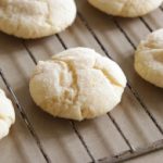 Several round, golden-brown cookies cool on a metal wire rack. The cookies have a slightly cracked surface and a soft, crumbly appearance.