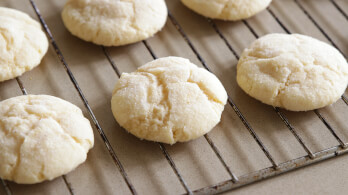 Several round, golden-brown cookies cool on a metal wire rack. The cookies have a slightly cracked surface and a soft, crumbly appearance.