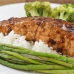 A plate of teriyaki-glazed salmon served on a bed of white rice, accompanied by steamed asparagus and broccoli florets. The dish is presented on a white plate, with the vegetables neatly arranged around the salmon.