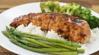 A plate of teriyaki-glazed salmon served on a bed of white rice, accompanied by steamed asparagus and broccoli florets. The dish is presented on a white plate, with the vegetables neatly arranged around the salmon.