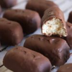 Rows of chocolate-covered rectangular candies are displayed on a wire rack. One candy is bitten to reveal a creamy, white filling inside.
