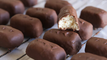 Rows of chocolate-covered rectangular candies are displayed on a wire rack. One candy is bitten to reveal a creamy, white filling inside.