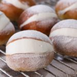 A batch of cream-filled doughnuts rests on a cooling rack. The doughnuts are covered with a light dusting of powdered sugar, and a generous amount of creamy filling is visible in the middle of each one.