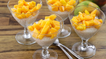 Four glass cups filled with creamy rice pudding topped with diced mango pieces are arranged on a wooden surface. Two silver spoons are placed between the cups. A blurred background hints at more mangoes.