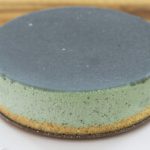 A close-up of a round cake sitting on a white plate. The cake has a smooth, dark green glaze on top and appears to have a green frosting layer with a light brown crust at the bottom. The background is a wooden surface.