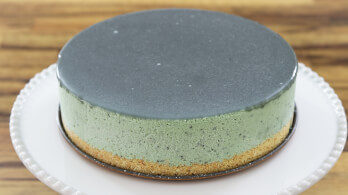 A close-up of a round cake sitting on a white plate. The cake has a smooth, dark green glaze on top and appears to have a green frosting layer with a light brown crust at the bottom. The background is a wooden surface.