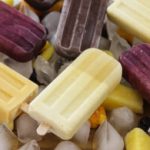 An assortment of colorful popsicles in yellow, purple, and chocolate brown rest atop ice cubes and scattered frozen fruits including mango and berries. The frozen treats are arranged in a visually appealing pattern, creating a refreshing, summery scene.